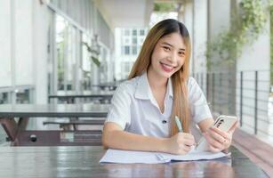 Young Asian woman student in uniform using smartphone and writing something about work. Documents on the table at university in Thailand. Her face with smiling in a working at to search information. photo