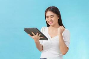 Cute Asian woman in white shirt is smiling happily and satisfy while look at on tablet in her hand isolated on blue background photo