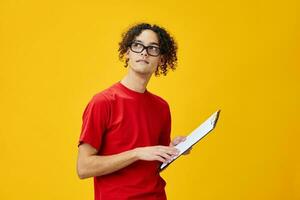 Thinking myopic young student man in red t-shirt funny eyewear holds tablet folder with study notes posing isolated on over yellow studio background. Free place for ad. Education College concept photo