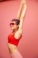 Fashion portrait of a woman with a short haircut in colored sunglasses with unusual accessories with earrings smiling on a pink bright background with a fitness body dancing photo