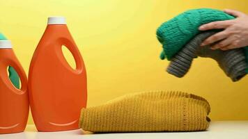 Stack of washed folded clothes and plastic orange large bottles with liquid detergent stand on a white table, yellow background. Routine homework video