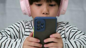 Cute elementary school girl wearing headphones holding a smartphone. Happy Asian girl studying online on smartphone or homeschooling, listening to music or playing games. video