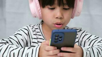 süß elementar Schule Mädchen tragen Kopfhörer halten ein Smartphone. glücklich asiatisch Mädchen studieren online auf Smartphone oder häuslicher Unterricht, Hören zu Musik- oder spielen Spiele. video