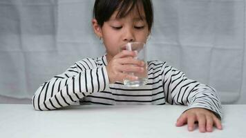 fofa pequeno menina bebendo água a partir de vidro em branco cortina fundo. saudável estilo de vida e refresco conceito. video