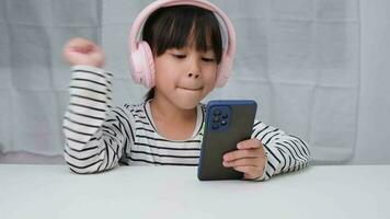 carino elementare scuola ragazza indossare cuffie Tenere un' smartphone. contento asiatico ragazza studiando in linea su smartphone o istruzione a casa, ascoltando per musica o giocando Giochi. video