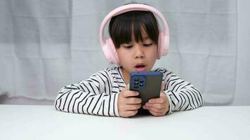 Cute elementary school girl wearing headphones holding a smartphone. Happy Asian girl studying online on smartphone or homeschooling, listening to music or playing games. video
