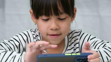 Cute elementary school girl wearing headphones holding a smartphone. Happy Asian girl studying online on smartphone or homeschooling, listening to music or playing games. video