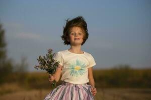 contento pequeño niña en el prado con un ramo de flores de flores un niño en un hermosa verano campo con desarrollando cabello. foto