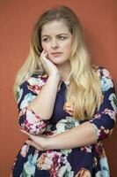 A sad blonde girl with blue eyes in a blue dress with a floral print stands near a burgundy wall. photo