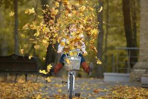 Young woman rides a bicycle in an autumn park under a bunch of falling leaves. Beautiful autumn background with girl and leaves. photo