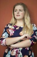 A pensive blonde girl with blue eyes in a blue dress with a floral print stands near a burgundy wall. photo