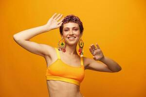 Portrait of a young woman with a short haircut and colored hair smiling in yellow and showing her tongue at the camera on an orange background dancing with earrings accessories in the studio photo