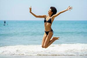 Black woman taking selfie while jumping near sea photo