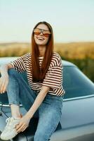 A fashionable woman smiles sweetly in stylish sunglasses, a striped t-shirt and jeans, sits on the trunk of a car and looks at the beautiful nature of autumn. Travel lifestyle photo