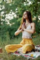 A young hippie woman meditates in nature in the park, sitting in a lotus position on her colorful plaid and enjoying harmony with the world in eco-clothing photo