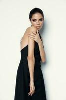 hermosa mujer modelo en un negro vestir toques sí misma con manos en un ligero antecedentes foto