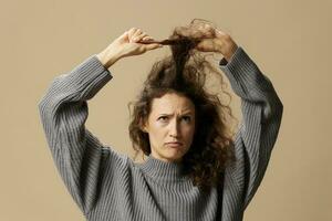 Funny thoughtful curly beautiful female in gray casual sweater with hairbrush comb pulls curls up posing isolated on over beige pastel background. Problematic unruly damaged hair concept. Copy spaceFunny thoughtful curly beautiful female in gray casual sw photo