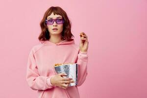 Cool movie. Confused cute redhead lady in pink hoodie sunglasses with popcorn posing isolated on over pink studio background show copy space Banner. Fashion Cinema concept. Entertainment offer photo