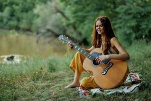 joven hippie mujer sentado al aire libre en un tartán y jugando su guitarra en el orilla del río dentro el puesta de sol foto