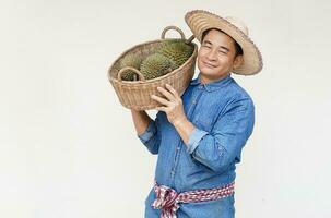 Handsome Asian man farmer wears hat, blue shirt, holds basket of durian fruits on shoulder. Concept, agriculture occupation. Thai farmers grow durian fruits as economic and export fruits production photo