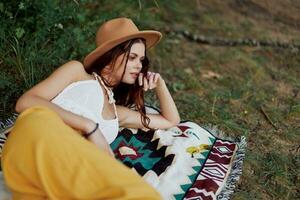 un mujer en Respetuoso del medio ambiente ropa en un hippie Mira es acostado en un de colores tartán sonriente y mirando a un otoño puesta de sol en naturaleza en el parque. estilo de vida en un viaje de armonía con el mundo foto