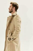 man holding hands in pockets of beige coat side view light background photo