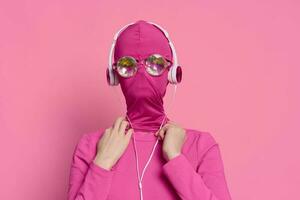 Art photo conceptual portrait of a woman in a pink mask covering her face with shiny glowing glasses and wearing headphones on a bright pink background holding wires with her hands