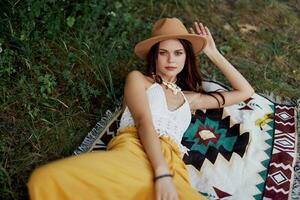 A woman in eco-friendly clothing in a hippie look is lying on a colored plaid smiling and looking at an autumn sunset in nature in the park. Lifestyle on a journey of harmony with the world photo