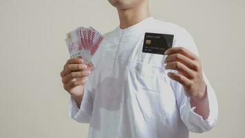 retrato de atractivo asiático musulmán hombre en blanco camisa con casquete participación uno cien mil rupia y presentación crédito tarjeta. aislado imagen en gris antecedentes foto