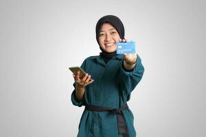 Portrait of cheerful Asian muslim woman with hijab, showing credit card while holding mobile phone. Financial and savings concept. Isolated image on white background photo