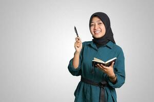 Portrait of excited Asian muslim woman with hijab writing on note book and pointing up with pen. Advertising concept. Isolated image on white background photo