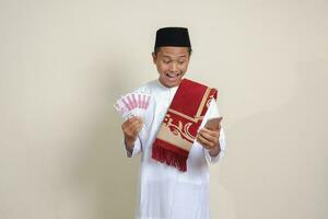 Portrait of attractive Asian muslim man in white shirt showing one hundred thousand rupiah while using mobile phone. Financial and savings concept. Isolated image on gray background photo