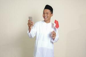 Portrait of attractive Asian muslim man in white shirt with skullcap holding indonesia flag while showing mobile phone. Isolated image on gray background photo