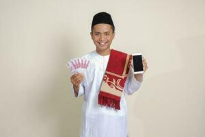 Portrait of attractive Asian muslim man in white shirt with skullcap showing one hundred thousand rupiah while showing blank screen mobile phone. Financial and shopping concept. Isolated image on gray photo