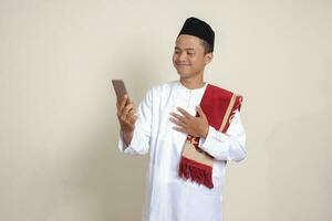 Portrait of attractive Asian muslim man in white shirt taking picture of himself or selfie, saying hi and waving his hand during video call. Isolated image on gray background photo