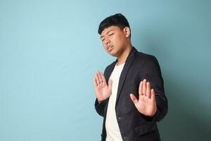 Portrait of young Asian business man in casual suit with rejecting hand gestures while looking at camera. Isolated image on blue background photo