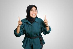 Portrait of excited Asian hijab woman in casual outfit standing against white background, showing product and pointing with hand and finger to the side. Advertising concept. photo