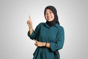 Portrait of excited Asian hijab woman in casual outfit standing against white background, showing product and pointing with hand and finger to the side. Advertising concept. photo