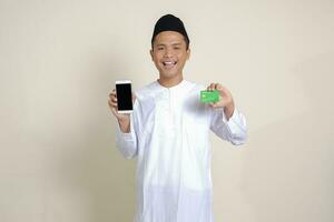 retrato de atractivo asiático musulmán hombre en blanco camisa con casquete participación un móvil teléfono y presentación crédito tarjeta. aislado imagen en gris antecedentes foto