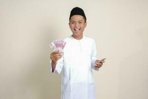 Portrait of attractive Asian muslim man in white shirt showing one hundred thousand rupiah while using mobile phone. Financial and savings concept. Isolated image on gray background photo