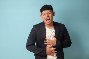 Portrait of young Asian business man in casual suit bursting out laugh while holding stomach with both hands. Isolated image on blue background photo