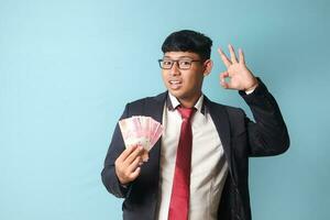 Portrait of young Asian business man in casual suit holding thousand rupiahs while making ok gestures. Isolated image on blue background photo