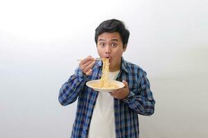 Portrait of happy Asian man in blue plaid shirt eating delicious instant noodles with chopsticks served on plate. Isolated image on white background photo