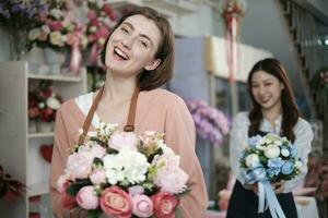 Young beautiful White female florist worker with bunch of blossoms and her colleague smile and look at camera at colorful flower shop in Valentine, small business occupation, happy SME entrepreneur. photo