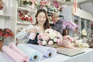 Young beautiful Asian female florist worker water spraying a flora bunch with old male employee, happy work in colorful flower shop store, fresh bloom bouquets decorating, SME business entrepreneur. photo