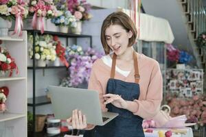 Portrait of one young White female florist owner in apron with laptop in bright and beautiful flower shop store smiles with online shopping jobs, small e-commerce business, happy SME entrepreneur. photo