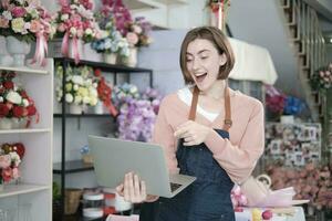 One young beautiful White female florist owner surprised and shocked purchase order profit, online income work in laptop at bright flower shop store, small e-commerce business, happy SME entrepreneur. photo