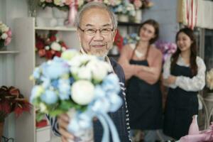 Senior Asian male florist owner in apron gives bunch of blossom smiles and looks at camera in front of her colleague's team in colorful flower shop, small business occupation, happy SME entrepreneur. photo