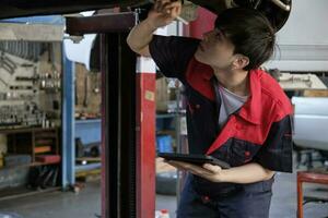 Professional young Asian male motor mechanic inspects undercarriage of electric car lifted by forklift jack for repair at garage, automotive maintenance service works industry occupation business. photo