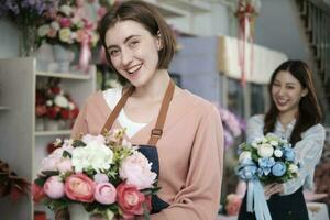 Young beautiful White female florist worker with bunch of blossoms and her colleague smile and look at camera at colorful flower shop in Valentine, small business occupation, happy SME entrepreneur. photo